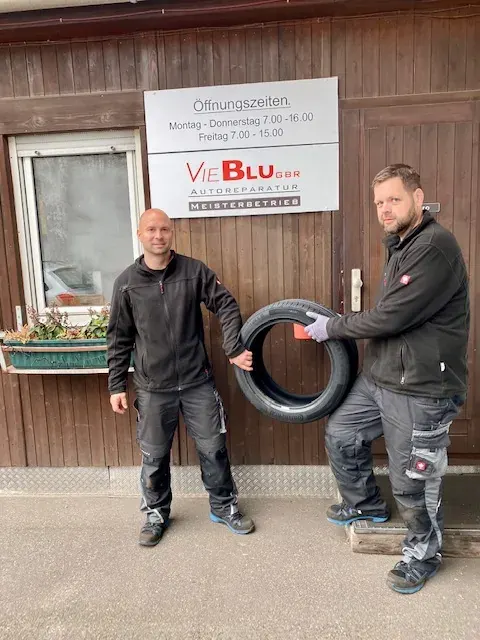 Zwei Mitarbeiter der VieBlu GbR mit einem Reifen vor dem Werkstatteingang unter dem Schild mit den Öffnungszeiten